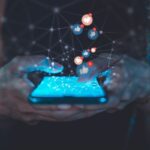 Hands holding a mobile phone. Icons featuring thumbs up and hearts are rising from the phone indicating social media engagement