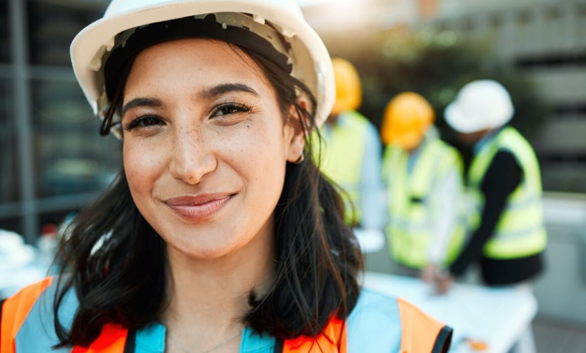 A construction worker, wearing a hard hat and hi-viz jacket smiles at the camera with three of her colleagues standing behind her.