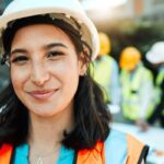 A construction worker, wearing a hard hat and hi-viz jacket smiles at the camera with three of her colleagues standing behind her.